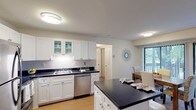 This single-floor residence offers a thoughtfully designed layout that balances functionality with comfort. The kitchen is a standout feature, equipped with crisp white shaker-style cabinetry — including glass-front upper cabinets — dark countertops, and a full suite of stainless steel appliances including a refrigerator, electric range with over-the-range microwave, and dishwasher. Light hardwood-style flooring anchors the kitchen and flows into the adjacent areas. A breakfast bar peninsula connects the kitchen to the living and dining spaces, making the layout feel open and cohesive. The dining area is brightened by a large sliding glass door with vertical blinds that invite natural light and provide access to an outdoor area. The living room features soft carpeting, neutral blue-grey walls, and a welcoming, relaxed atmosphere. The bedroom is generously proportioned, with soft carpet, sage-toned accent walls, ample room for a full furniture arrangement, and a window that brings in natural light. The full bathroom is clean and well-organized, featuring a single vanity with white cabinetry and a tub-shower combination. Throughout the residence, a calm, neutral color palette and consistent finishes create a harmonious and livable environment.
