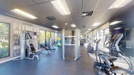 This modern fitness center is designed to support a full range of workout routines within a bright, open layout. The space features durable rubber flooring throughout, providing a safe and functional surface for exercise. A dropped ceiling with recessed panel lighting ensures consistent, even illumination across the entire facility.

Large floor-to-ceiling windows line multiple walls, flooding the interior with natural light and offering views of an outdoor pool area, landscaped grounds, and surrounding greenery. These expansive windows create an open, airy atmosphere that enhances the overall workout experience.

The cardio zone is well-appointed with treadmills, elliptical trainers, stationary bikes, and stair-climbing machines, all positioned to take advantage of the outdoor views. The strength training area includes cable machines, a multi-functional power rack, adjustable benches, and a full dumbbell rack, supported by large wall mirrors that allow users to monitor their form. Stability balls and other functional training accessories are also available throughout the space.

A standout design feature is the motivational typography wall mural, which displays words and phrases such as "Fitness," "Focus," "Believe," "Transformation," and "Never Give Up," reinforcing an energizing and goal-oriented atmosphere. At least one mounted flat-screen television is also present. The overall design is clean, modern, and purposefully organized to maximize usability.