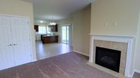 This single-floor home is designed around an open-concept floor plan that seamlessly connects the living and kitchen areas, creating a spacious and functional layout. The living area features wall-to-wall carpeting in a neutral tone, providing a comfortable and inviting atmosphere. A tile-surround fireplace with a classic white mantel serves as a focal point, adding warmth and character to the space.

The kitchen is finished with large-format tile flooring and features rich, dark wood cabinetry that contrasts elegantly with light-colored countertops. A central island offers additional workspace and casual seating potential. Appliances are integrated into the cabinetry layout, and a window above the sink allows natural light to brighten the workspace.

An elegant chandelier over the kitchen area adds a decorative touch, complementing the overall warm, neutral color palette found throughout the home. Glass-panel doors lead directly to the outdoors, flooding the space with natural light and providing easy access to the exterior. The combination of carpet and tile flooring clearly delineates the living and kitchen zones while maintaining a cohesive aesthetic throughout.