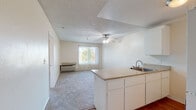 This single-story unit in Martinez, California offers a practical, open layout designed for comfortable everyday living. The living area is generously sized, featuring soft wall-to-wall carpeting, a ceiling fan with light fixture, textured ceiling, and a large sliding glass window dressed with vertical blinds that invite natural light into the space. A wall-mounted HVAC unit provides climate control directly in the living area.

The kitchen is positioned in an open-concept arrangement, separated from the living space by a peninsula counter with a light-toned laminate surface. White cabinetry lines the kitchen walls, complemented by a stainless steel sink, an electric range with overhead ventilation, and a full-size white refrigerator. The kitchen floor features warm hardwood-style flooring, contrasting with the carpeted living and bedroom areas.

The bedroom is a quiet, well-proportioned room with plush carpeting, a window with vertical blinds, and a textured ceiling. The bathroom includes a tub/shower combination with accessibility grab bars, wood-style flooring, a vanity with a light stone-finish countertop, and clean white tile surround, offering a functional and accessible design.