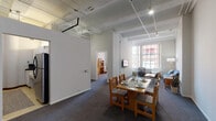 Located at 731 S Plymouth in Chicago's South Loop neighborhood, this single-floor unit offers a thoughtfully arranged layout with a variety of functional spaces. The open common area combines a dining zone — anchored by a wood dining table with seating for several — and a comfortable living room featuring a wall-mounted TV, a blue sofa, armchairs, and a coffee table. Large grid-pane windows flood the living area with natural light and frame views of the surrounding Chicago cityscape.

The bedrooms vary in character: some feature soaring ceilings with exposed structural details and transom windows that enhance the sense of height and openness, while others offer a more intimate feel with ceiling fans, individual wardrobes, dressers, and desks. All bedrooms are carpeted and benefit from natural light through sizable windows with city views.

The galley kitchen is efficiently designed with white cabinetry lining both walls, ample counter space, a stainless steel sink, and standard appliances. The bathroom is clean and straightforward, featuring a wide vanity sink, wall-mounted mirror with Hollywood-style lighting, and a shower with curtain. Throughout the unit, gray carpeting, neutral walls, and track lighting create a consistent, functional aesthetic suited to shared or transitional living.