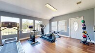 This fitness center in Austin, TX offers a bright and functional single-room exercise space designed to support a well-rounded workout routine. The room features warm hardwood flooring throughout, providing a clean and durable surface suited for a variety of exercise activities. Vaulted ceilings lend an airy, open feel to the space, while multiple windows along the walls allow ample natural light to fill the room and offer views of the surrounding greenery outdoors.

The exercise room is equipped with a diverse selection of cardio and strength equipment, including a treadmill, elliptical trainer, stationary bike, and a rowing machine — all positioned on protective floor mats. A freestanding punching bag adds a boxing and combat fitness option, while resistance bands mounted on the wall provide supplementary strength training versatility. A workout poster and posted fitness center rules contribute to an organized, informative environment.

Access to the space is provided through a decorative white entry door with an oval glass insert, as well as French doors that connect to an adjacent area. The cool gray-toned walls complement the natural wood floors, creating a calm and motivating atmosphere. Overhead fluorescent lighting ensures the space remains well-lit at all times. This fitness center delivers a comprehensive and welcoming workout environment within a compact, efficiently utilized footprint.