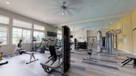 This single-floor fitness center is thoughtfully designed to support a comprehensive workout experience. The space is generously sized, with an open layout that accommodates a diverse range of exercise equipment without feeling crowded. Light-toned, striped hardwood-style flooring runs throughout, contributing to a clean and modern aesthetic.

The cardio zone is well-appointed with multiple treadmills, elliptical machines, a stationary bike, and a rowing machine, all positioned near a bank of large windows that allow abundant natural light and outdoor views. Wall-mounted televisions are strategically placed throughout the space to keep users entertained during workouts.

The strength training area includes a cable machine, a leg press, adjustable benches, a dumbbell rack, and a selection of medicine balls and exercise balls. A full-length mirror wall visually expands the space while allowing users to monitor their form. An additional motivational mural in bold yellow with fitness-themed typography adds energy and character to one accent wall.

Ceiling fans with integrated lighting and recessed fixtures provide comfortable air circulation and even illumination throughout. The overall ambiance is bright, clean, and energizing, making this fitness center a functional and inviting space for exercise.