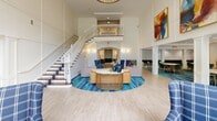 Located at 1800 Palmer Rd in Fort Washington, Maryland, this expansive single-floor community space offers an impressive and versatile layout designed for both relaxation and social engagement. The centerpiece is a dramatic two-story lobby anchored by soaring ceilings, a grand staircase with white balusters and wood-toned treads, and an open upper-level mezzanine balcony that overlooks the main gathering area below. Bold abstract artwork adorns the walls, adding vibrant pops of color that complement the blue-and-neutral coastal-inspired palette carried throughout the space. The living area features oversized windows stretching nearly floor to ceiling, dressed in elegant two-tone navy and cream curtains that frame lush outdoor views and flood the interior with natural light. Comfortable seating arrangements—including leather sofas and plaid upholstered armchairs—create inviting conversation nooks. A dedicated dining area offers round tables with modern teal upholstered chairs set atop a rich blue patterned area rug, alongside a bar or refreshment counter with navy cabinetry. A game room with a billiards table provides additional recreational options. Light wood-look flooring flows seamlessly throughout, unifying the open, airy floor plan and reinforcing the welcoming, resort-like ambiance of this well-appointed community hub.