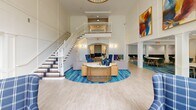 Located at 1800 Palmer Rd in Fort Washington, Maryland, this expansive single-floor community space offers an impressive and versatile layout designed for both relaxation and social engagement. The centerpiece is a dramatic two-story lobby anchored by soaring ceilings, a grand staircase with white balusters and wood-toned treads, and an open upper-level mezzanine balcony that overlooks the main gathering area below. Bold abstract artwork adorns the walls, adding vibrant pops of color that complement the blue-and-neutral coastal-inspired palette carried throughout the space. The living area features oversized windows stretching nearly floor to ceiling, dressed in elegant two-tone navy and cream curtains that frame lush outdoor views and flood the interior with natural light. Comfortable seating arrangements—including leather sofas and plaid upholstered armchairs—create inviting conversation nooks. A dedicated dining area offers round tables with modern teal upholstered chairs set atop a rich blue patterned area rug, alongside a bar or refreshment counter with navy cabinetry. A game room with a billiards table provides additional recreational options. Light wood-look flooring flows seamlessly throughout, unifying the open, airy floor plan and reinforcing the welcoming, resort-like ambiance of this well-appointed community hub.