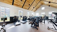 Located at 1 Samuel Peterson Dr in Titusville, NJ, this fitness center is a thoughtfully designed, single-floor facility that offers a generous workout space across two distinct areas. The main gym floor immediately impresses with its dramatic vaulted ceiling clad in warm natural wood, supported by exposed black structural beams and illuminated by pendant lighting and a ceiling fan. Tall windows line the walls on multiple sides, flooding the interior with natural light and offering views of the surrounding community. The two-tone wall treatment — crisp white above and deep charcoal below — lends a modern, polished aesthetic throughout. The primary workout area is comprehensively equipped with a full lineup of TRUE-brand treadmills, ellipticals, rowing machines, stair climbers, and cycling bikes, alongside a robust selection of selectorized strength machines and a full dumbbell rack with battle ropes. Large mirrors maximize the sense of space and support proper form during training. A secondary room, finished with light wood flooring, provides a more intimate setting ideal for stretching, yoga, or supplemental workouts. The durable rubber-speckled flooring throughout the main gym ensures both safety and comfort. Overall, the facility combines functional versatility with an upscale, airy design.