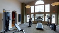 This fitness center is designed to offer a functional and comfortable workout environment. The space is defined by its impressively high ceilings, which create an open, airy atmosphere that enhances the overall experience. A large arched window wall dominates one side of the room, allowing abundant natural light to pour in and providing views of the outdoor surroundings — making workouts feel less confined and more energizing.

The room features a mix of cardio and strength training equipment, including a treadmill, a recumbent stationary bike, and an adjustable weight bench, along with additional strength training apparatus. The flooring consists of durable, low-pile carpet well-suited for a fitness setting. A prominent pendant light fixture hangs from the ceiling, complementing the natural light with warm overhead illumination.

The walls are finished in a neutral beige tone with contrasting dark green accent walls, adding a sense of depth and visual interest to the space. A glass-paneled door provides direct exterior access, further reinforcing the connection to the outdoors. Safety features such as clearly marked emergency exit signage are also present. Overall, the fitness center balances practicality with a bright, welcoming ambiance suited for a variety of workout routines.