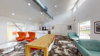 Located at 8787 Hammerly Blvd in Houston, Texas, this single-story community clubhouse offers a lively and well-appointed gathering space designed for both socializing and relaxation. The main area features soaring ceilings with exposed industrial-style ductwork, adding an urban, contemporary character to the bright, white-walled interior. Natural light streams in through clerestory windows positioned high along the walls, further enhancing the airy, open feel of the space.

The central gathering area is anchored by a solid wood communal dining table surrounded by a mix of modern seating, including iconic egg chairs in bold orange and classic molded chairs in white and red, all set atop a patterned, multi-tonal carpet. Yellow pendant lights add a cheerful accent overhead.

A dedicated lounge area features retro-inspired teal sofas arranged around a marble-topped coffee table, with a large picture window offering direct views of an outdoor patio, pool, and lush palm trees beyond — creating a seamless indoor-outdoor connection.

The kitchen area is fully equipped with stainless steel appliances including a French door refrigerator and a coffee station, complemented by granite countertops, white upper cabinetry, and wood-look tile flooring — providing all the amenities needed for community gatherings and events.