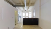 This single-floor space showcases a compelling blend of industrial and contemporary design elements. The most striking architectural feature is the exposed concrete ceiling, complete with raw beams and a large silver ventilation duct running its length, lending an authentic loft character to the interior. Polished light-toned flooring reflects natural light throughout, enhancing the sense of openness and continuity across the space.

The open-concept kitchen serves as a focal point, anchored by a substantial island with sleek dark cabinetry and a light countertop that provides a strong visual contrast against the bright surroundings. Adjacent kitchen cabinetry continues the dark-and-light palette, with stainless steel appliances visible in the background. Track lighting runs along the ceiling, supplementing the generous natural light that pours in through a row of large windows at the far end of the space.

The layout flows seamlessly, with white walls creating a clean, modern backdrop that balances the rawness of the industrial ceiling elements. A doorway leads to an additional room, suggesting functional versatility. Overall, the space communicates an urban, sophisticated ambiance well-suited to modern living or working.