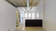 This single-floor space showcases a compelling blend of industrial and contemporary design elements. The most striking architectural feature is the exposed concrete ceiling, complete with raw beams and a large silver ventilation duct running its length, lending an authentic loft character to the interior. Polished light-toned flooring reflects natural light throughout, enhancing the sense of openness and continuity across the space.

The open-concept kitchen serves as a focal point, anchored by a substantial island with sleek dark cabinetry and a light countertop that provides a strong visual contrast against the bright surroundings. Adjacent kitchen cabinetry continues the dark-and-light palette, with stainless steel appliances visible in the background. Track lighting runs along the ceiling, supplementing the generous natural light that pours in through a row of large windows at the far end of the space.

The layout flows seamlessly, with white walls creating a clean, modern backdrop that balances the rawness of the industrial ceiling elements. A doorway leads to an additional room, suggesting functional versatility. Overall, the space communicates an urban, sophisticated ambiance well-suited to modern living or working.