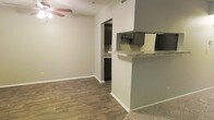 This single-story space showcases an open-concept design that seamlessly connects the living area and kitchen. The living area features wood-style plank flooring, neutral beige walls, and a ceiling fan, creating a comfortable and functional environment. A partial wall with a granite-topped breakfast bar serves as the dividing element between the living space and kitchen, providing both a visual boundary and a casual dining or workspace surface. The kitchen, visible through an arched passageway, is equipped with dark cabinetry offering ample storage and dark appliances that contrast cleanly against the lighter countertops and walls. The transition from wood-style flooring in the main living area to carpeted flooring hints at the layout's thoughtful zoning of spaces. The overall color palette is kept neutral with soft beige and gray tones, contributing to a clean, move-in-ready aesthetic. The open sightlines between the kitchen and living area enhance the sense of space, making the layout practical for everyday living and entertaining.