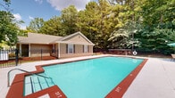 Located at 2319 College Dr in Phenix City, Alabama, this outdoor pool facility offers a well-maintained recreational space ideal for leisure and relaxation. The centerpiece is a generously sized swimming pool with clearly marked depth indicators ranging from 3 feet to 5 feet, providing safe and accessible swimming for a range of users. The pool features built-in entry steps and metal handrails for easy access.

Surrounding the pool is a spacious concrete deck finished with distinctive red-bordered edges, offering ample room for lounging and outdoor seating. A patio umbrella and seating area are positioned along the deck, providing a shaded retreat. A life ring is mounted near the pool, reflecting attention to safety.

Adjacent to the pool is a single-story pool house with a gabled roof, tan siding, and shuttered windows, which likely serves as a utility or support facility for the pool area. The entire space is enclosed by a black wrought iron fence, ensuring security while maintaining an open, airy feel.

The backdrop of the space is defined by a lush canopy of mature pine and deciduous trees, creating a serene, natural atmosphere. Abundant natural light and the vibrant greenery of the surrounding landscape contribute to a refreshing, resort-like ambiance throughout the outdoor space.