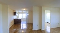 This single-floor space showcases a well-connected layout where the living area flows naturally into the kitchen, creating an open and functional floor plan. The living area is defined by rich, polished parquet wood flooring that adds warmth and character, reflecting ambient light to enhance the sense of brightness throughout the space.

A structural column serves as a subtle divider between the living zone and the kitchen area, adding architectural interest while maintaining the open feel. The kitchen features dark-toned cabinetry with a contrasting light countertop and includes a full-sized white refrigerator. Tile flooring in the kitchen provides a practical transition from the parquet in the adjoining living space. A large window in the kitchen allows generous natural light to fill the area, contributing to the overall airy atmosphere.

The walls are finished in a clean, neutral cream tone throughout, complementing both the dark cabinetry and the warm wood flooring. Flush-mount ceiling light fixtures provide consistent overhead lighting. A doorway visible to the right suggests additional rooms beyond, further extending the functionality of this straightforward, well-lit single-floor layout.