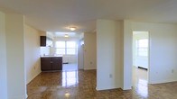 This single-floor space showcases a well-connected layout where the living area flows naturally into the kitchen, creating an open and functional floor plan. The living area is defined by rich, polished parquet wood flooring that adds warmth and character, reflecting ambient light to enhance the sense of brightness throughout the space.

A structural column serves as a subtle divider between the living zone and the kitchen area, adding architectural interest while maintaining the open feel. The kitchen features dark-toned cabinetry with a contrasting light countertop and includes a full-sized white refrigerator. Tile flooring in the kitchen provides a practical transition from the parquet in the adjoining living space. A large window in the kitchen allows generous natural light to fill the area, contributing to the overall airy atmosphere.

The walls are finished in a clean, neutral cream tone throughout, complementing both the dark cabinetry and the warm wood flooring. Flush-mount ceiling light fixtures provide consistent overhead lighting. A doorway visible to the right suggests additional rooms beyond, further extending the functionality of this straightforward, well-lit single-floor layout.