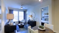 This well-designed single-floor residence offers a cohesive and elegantly styled interior that balances comfort with modern aesthetics. The open-concept layout allows the living, dining, and sleeping areas to flow naturally into one another, creating a sense of spaciousness and connectivity throughout the unit.

The living area is anchored by rich, dark hardwood flooring and features a ceiling fan with integrated lighting. Large windows dressed with navy blue curtains invite an abundance of natural light while adding a bold contrast to the neutral walls. The space is furnished with a plush sofa, a tufted accent chair, and a round coffee table atop a patterned blue area rug, lending a curated, contemporary feel.

A striking sunburst mirror adorns the wall above a sleek black credenza, serving as a focal point that ties the living and dining areas together. The dining area features a round wood table surrounded by cream upholstered chairs, creating an inviting space for meals and gatherings.

Adjacent to the main living area, the bedroom is visible through an open doorway, continuing the same refined palette with coordinated furnishings, a ceiling fan, and a decorative round mirror. The consistent design language throughout the space reflects a thoughtful, detail-oriented approach to interior styling.