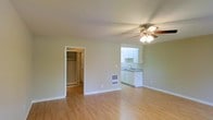This single-floor space offers a clean, open layout designed for comfortable everyday living. The main living area is finished with warm light wood laminate flooring that runs throughout, lending a cohesive and inviting feel to the space. A ceiling fan with an integrated light fixture is centrally positioned overhead, providing both airflow and ambient illumination.

The kitchenette is seamlessly integrated into the living area, featuring white cabinetry, a countertop with a built-in sink, and an overhead cabinet for additional storage. The white finishes of the kitchen create a bright contrast against the soft, neutral-toned walls that envelop the entire space.

A generously sized closet is accessible directly from the main room, offering ample storage with a visible hanging rod and shelving. A floor-level wall vent is also present, indicating a built-in heating and ventilation system for year-round comfort.

The walls throughout are painted in a soft, muted green-white tone that keeps the space feeling light and airy. The straightforward, functional design of the space makes it highly adaptable, with clean lines and minimal ornamentation that allow for flexible furnishing arrangements. Overall, the space presents a practical and well-maintained environment with thoughtful built-in features.