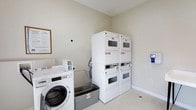 This shared laundry facility is a clean, efficiently organized space designed to accommodate the laundry needs of its users. The room features neutral-toned walls and light flooring, creating a bright and tidy environment. Multiple front-loading washing machines are positioned along one wall, each equipped with coin-operated or card payment controls for convenient use. Opposite, two sets of stacked dryers provide ample drying capacity, maximizing the use of vertical space. A utility basin sits between the machines, offering a practical solution for hand-washing or pre-soaking garments. A card payment terminal mounted on the wall provides a modern, cashless payment option. Posted instructions and signage ensure users can operate the machines with ease. The space is straightforward in its design, prioritizing function and accessibility. Clean baseboards and well-maintained equipment reflect the care taken in upkeeping the facility, making it a reliable and user-friendly laundry room.