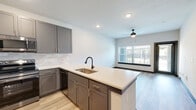 Located at 21447 FM-529 in Katy, TX, this single-floor unit offers a clean, contemporary design with a cohesive neutral color palette and light wood-look flooring that flows seamlessly throughout the space. The open-concept kitchen is a standout feature, showcasing gray shaker-style cabinetry, light quartz countertops, a subway tile backsplash, and a full suite of stainless steel appliances including a range, over-the-range microwave, dishwasher, and refrigerator. A kitchen island with an integrated sink and matte black faucet provides additional prep space while connecting the kitchen to the living area. The living space is bright and open, benefiting from natural light through a large window fitted with blinds, complemented by a ceiling fan overhead. The bedroom continues the airy feel with its own window, ceiling fan, and light hardwood-style flooring, offering a comfortable and well-lit retreat. The bathroom is finished with clean white walls, a soaking tub with shower combination, curved curtain rod, and dark-accented panel doors that echo the modern aesthetic found throughout. Recessed lighting and dark baseboards add polished finishing touches to this well-designed unit.
