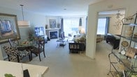 This single-floor residence offers an open, flowing layout that connects multiple living areas seamlessly. The main living room is anchored by a classic fireplace framed with a painted mantel, above which decorative artwork adds a personalized touch. Large windows dressed with elegant drape panels allow abundant natural light to fill the space, creating a bright and welcoming atmosphere. The room is finished with wall-to-wall carpeting in a neutral tone, contributing to a warm and comfortable feel throughout.

A dining area is positioned to one side, furnished with a dark wood table and matching chairs, providing a defined yet integrated space for meals. The living area accommodates a comfortable sofa arrangement centered around a glass-top coffee table, complemented by a bookcase unit that adds both function and character. An open archway leads into an adjacent room, likely a sunroom or secondary sitting area, where additional natural light pours in through bright windows. The neutral color palette of whites and soft grays throughout the space creates a cohesive, understated aesthetic suitable for a variety of décor styles.