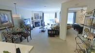 This single-floor residence offers an open, flowing layout that connects multiple living areas seamlessly. The main living room is anchored by a classic fireplace framed with a painted mantel, above which decorative artwork adds a personalized touch. Large windows dressed with elegant drape panels allow abundant natural light to fill the space, creating a bright and welcoming atmosphere. The room is finished with wall-to-wall carpeting in a neutral tone, contributing to a warm and comfortable feel throughout.

A dining area is positioned to one side, furnished with a dark wood table and matching chairs, providing a defined yet integrated space for meals. The living area accommodates a comfortable sofa arrangement centered around a glass-top coffee table, complemented by a bookcase unit that adds both function and character. An open archway leads into an adjacent room, likely a sunroom or secondary sitting area, where additional natural light pours in through bright windows. The neutral color palette of whites and soft grays throughout the space creates a cohesive, understated aesthetic suitable for a variety of décor styles.