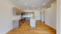 This single-floor unit is designed with a clean, contemporary aesthetic and an efficient open-concept layout. The kitchen serves as the centerpiece of the main living area, outfitted with warm-toned cabinetry extending to the upper walls for ample storage, light granite countertops, and a full suite of stainless steel appliances including a range, microwave, dishwasher, and refrigerator. A central island with a matching granite surface provides additional workspace and casual seating, while light hardwood flooring ties the kitchen and living area together seamlessly. Recessed lighting throughout the open space ensures consistent, even illumination. The living area extends toward a set of French doors that open to a private balcony, bringing in natural light and an outdoor connection. The bedroom is carpeted for comfort and features a large window framing views of the surrounding urban streetscape. The bathroom includes a full-size soaking tub with a shower, clean white surround, and chrome fixtures. Overall, the unit presents a bright, move-in-ready environment with a cohesive modern palette of neutral tones and quality finishes.