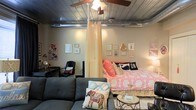 This single-floor studio showcases a distinctive industrial-chic aesthetic, anchored by a standout metallic ceiling with exposed ductwork that adds raw architectural character to the space. A ceiling fan with integrated lighting serves as a functional centerpiece overhead.

The open-plan layout is thoughtfully divided into distinct zones without the use of permanent walls. A sheer curtain panel elegantly separates the sleeping area from the living space, offering a soft, airy sense of privacy. The sleeping zone features a full-size bed dressed in vibrant coral and pink bedding, flanked by a curated gallery wall that adds personality and warmth. A dedicated workspace with a desk and chair is tucked neatly into one corner.

The living area hosts a spacious grey sectional sofa paired with a glass-top coffee table and a patterned area rug, creating a comfortable and inviting lounge space. A classic Eames-style lounge chair and ottoman add a touch of mid-century modern sophistication.

Toward the back, a kitchen with a breakfast bar and pendant lighting provides a polished transition into the culinary space, complete with bar-height seating. Dark hardwood-style flooring runs throughout, tying all the zones together cohesively. The overall ambiance balances urban industrial elements with warm, personalized décor.