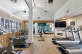 Located at 2052 Wilkins Ave in Napa, CA, this community amenity space features a thoughtfully designed fitness center complemented by a spacious outdoor pool area. The fitness center is housed within a vaulted-ceiling room that creates an airy, open atmosphere, with soaring cathedral-style ceilings and exposed white structural beams adding architectural character. Warm wood-tone flooring runs throughout, while horizontal wood-paneled wainscoting along the lower walls lends a contemporary, inviting aesthetic. Stylish pendant lighting, including woven rattan and globe-style fixtures, provides warm illumination throughout the space. The fitness floor is equipped with a comprehensive selection of cardio machines, including treadmills, ellipticals, and stair climbers, along with a free weight rack and a cable resistance machine. Full-length mirrors on a teal-painted accent wall visually expand the space and allow for form monitoring during workouts. A wall-mounted flat-screen television is positioned above built-in shelving for convenience during cardio sessions. A connected anteroom features bold teal walls, a geometric-patterned floor, and a modern chandelier. The outdoor pool area features a large, sparkling swimming pool surrounded by a generous sun deck furnished with lounge chairs, set against the backdrop of the surrounding residential buildings and mature trees.