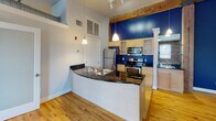 Located at 214 N Dunlop St in Petersburg, VA, this single-floor space blends historic character with modern updates. Exposed red brick walls are a defining architectural feature throughout, lending an industrial-chic aesthetic that is complemented by warm hardwood flooring found in the main living and open areas. Natural light flows generously through multiple double-hung windows, illuminating the spacious, open floor plan. The kitchen is a functional centerpiece, featuring stainless steel appliances, a stove and refrigerator, light wood cabinetry, dark granite countertops, and a curved peninsula with a built-in sink. A bold blue accent wall, pendant lighting, exposed wood ceiling beams, and decorative columns add visual interest and character to this area. Frosted glass partition doors connect the open living space to additional rooms, offering flexibility in use. The secondary rooms continue the exposed brick aesthetic with hardwood floors and windows providing outdoor views. The bathroom is well-appointed with tile flooring, a dark granite vanity, a large mirror, and a shower-tub combination. Exposed ductwork and ceiling fans are practical elements that enhance the loft-like industrial ambiance of this unique Petersburg property.