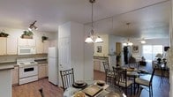 This single-floor residence offers a thoughtfully designed open-concept layout that connects the dining area and kitchen in a cohesive, functional arrangement. The dining space is anchored by a round glass-top table surrounded by ornate wrought-iron chairs, positioned beneath a multi-bulb chandelier with frosted glass shades that provides warm, focused illumination. A striking floor-to-ceiling mirror panel visually expands the space, reflecting both the dining and kitchen areas to create a sense of added depth and openness. Rich hardwood flooring runs throughout, lending warmth and continuity between spaces. The kitchen features white cabinetry paired with a matching refrigerator, a stainless steel sink, and a dishwasher, all arranged efficiently along the perimeter. A partial wall separates the kitchen from the dining area while maintaining visual connectivity. Additional pendant lighting echoes the dining fixture, reinforcing a cohesive design aesthetic throughout. White interior doors with paneled detailing contribute to the clean, classic character of the space. Greenery accents, placed atop the cabinetry, add a subtle natural touch to the otherwise neutral, light-toned interior palette.