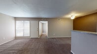 This single-floor residence offers a practical and well-organized layout designed for comfortable everyday living. The spacious living area is carpeted in warm, neutral tones and benefits from natural light streaming through blinded windows. A textured ceiling adds subtle character, while a ceiling-mounted light fixture provides ambient illumination. The living space connects seamlessly to a galley-style kitchen, separated by a countertop bar that provides both a visual boundary and additional workspace. The kitchen features warm honey-toned wood cabinetry, wood-look flooring, a double sink, and black appliances including a refrigerator, electric range, and dishwasher, all set beneath flush ceiling lighting. The bedrooms are generously sized with plush carpeting, neutral walls, and windows that invite natural light. One bedroom includes a large walk-in closet with built-in shelving, offering ample storage. The bathrooms are clean and functional, each featuring wood-look flooring, a vanity with granite-style countertops, a warm wood cabinet base, and a large mirror. The full bathroom includes a tub-shower combination with white tile surround. Throughout the home, a consistent neutral color palette and practical finishes create a cohesive and move-in-ready atmosphere.