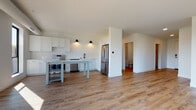 This single-floor unit offers a clean, contemporary design with a cohesive neutral palette throughout. The open-concept kitchen and living area serves as the heart of the space, featuring white shaker cabinetry, a subway tile backsplash, light quartz countertops, and a full suite of stainless steel appliances including an electric range, dishwasher, and French door refrigerator. A freestanding kitchen island with built-in shelving provides additional prep space and storage. Warm wood-tone luxury vinyl plank flooring runs through the main living area, complemented by stylish wall-mounted sconce lighting that adds an upscale touch. The spacious living room benefits from large, dark-framed windows that fill the room with natural light and offer elevated views of the surrounding cityscape and tree canopy. The bedroom, finished with plush carpet, also features a generous picture window with outdoor views. Two bathrooms serve the unit — one appointed with a soaking tub, white subway tile surround, and matte black fixtures, and a second with a walk-in tile shower. A dedicated in-unit laundry area houses a stacked Samsung washer and dryer. The overall design emphasizes brightness, functionality, and modern finishes throughout.