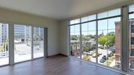 This single-floor space is defined by its striking floor-to-ceiling glass curtain wall, which spans nearly the entire exterior-facing side of the main living area. The expansive glazing floods the interior with abundant natural light throughout the day, creating an open, airy atmosphere that feels connected to the surrounding urban environment. The panoramic views take in a lively cityscape, including tree-lined streets, neighboring buildings of varied architectural styles, and a notable historic church steeple visible in the distance.

The living area features smooth, light-toned hardwood flooring that complements the neutral, cream-colored walls and contributes to a clean, contemporary aesthetic. Sliding glass doors positioned along one wall provide direct access to an outdoor balcony, further extending the sense of openness and allowing for fresh air and additional outdoor views of the streetscape below.

The space benefits from high ceilings that enhance the feeling of volume and spaciousness throughout the interior. Overall, the unit presents a modern, minimalist character with an emphasis on natural light, urban connectivity, and thoughtful architectural detailing.