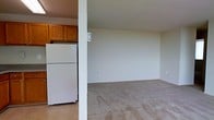 This single-floor residential space presents a straightforward and functional layout designed for comfortable everyday living. The kitchen is equipped with warm-toned wooden cabinetry that provides ample storage, complemented by a full-size white refrigerator and light-toned flooring that gives the area a clean, practical feel. A structural column subtly divides the kitchen from the adjacent living area, maintaining an open and connected atmosphere throughout the space. The living area is generously sized, featuring wall-to-wall carpeting in a neutral beige tone that adds warmth underfoot. The walls are finished in a soft, neutral gray-white palette, creating a calm and versatile backdrop that allows for a variety of interior styles and furnishing choices. The open flow between the kitchen and living spaces makes the layout feel cohesive and easy to navigate. Overall, this space offers a no-frills, move-in-ready environment with a clean and understated aesthetic, well-suited for those seeking a functional and adaptable living space on a single level.