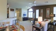 This two-floor residence is designed with an open and airy layout that maximizes both space and natural light. The main living area is a standout, featuring vaulted ceilings that lend a sense of grandeur, anchored by a ceiling fan and complemented by a striking arched window that floods the room with abundant daylight. The living room is finished with plush carpeting and offers ample space for comfortable seating arrangements, with a dedicated media console area along one wall. A bold accent wall adds visual depth and contrast to the otherwise light and neutral color palette. The dining area flows naturally from the living space, featuring bar-height seating that connects directly to the kitchen. The kitchen is partially open to the main living area via a raised counter, allowing for easy interaction between spaces. Throughout the main level, the layout demonstrates a thoughtful balance between open communal areas and defined functional zones. Windows fitted with blinds throughout allow for flexible light control while maintaining privacy. The overall ambiance of the space is warm and inviting, combining modern design elements with practical, livable features across both floors.