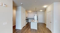 This single-floor residence showcases a thoughtfully designed open-concept layout that seamlessly connects the kitchen and living areas. The kitchen is a standout feature, equipped with stainless steel appliances, including a refrigerator and range, complemented by white cabinetry that extends to the upper walls. A central island with a light granite countertop and undermount sink serves as both a functional workspace and a natural gathering point, accented by pendant lighting overhead.

The countertops throughout the kitchen carry a consistent granite aesthetic, while the white cabinetry provides a clean, contemporary contrast. Rich hardwood-style flooring runs throughout the visible areas, adding warmth and continuity to the space. Recessed and flush-mount ceiling fixtures provide ample ambient lighting, supplemented by the pendant lights above the island.

The open layout allows for a natural flow between the kitchen and the adjacent living area, with neutral-toned walls enhancing the bright, airy atmosphere. A white paneled door is visible toward the rear of the space, suggesting access to additional rooms. The overall design balances modern functionality with a welcoming, residential aesthetic.