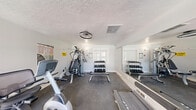 Located at 3135 Comanche Rd NE in Albuquerque, New Mexico, this dedicated fitness room offers a compact yet efficiently organized space for a well-rounded workout experience. The room features light gray walls, a textured ceiling with a ceiling fan for air circulation, and durable dark flooring designed to withstand heavy equipment use.

Natural light fills the space through a large sliding glass door and multiple windows fitted with blinds, providing views of an outdoor pool area and surrounding greenery. A full-wall mirror on one side enhances the sense of openness and allows users to monitor their form during exercise.

The fitness room is outfitted with a variety of cardio equipment, including a treadmill, elliptical trainer, and recumbent stationary bike. A multi-station cable weight machine anchors one corner of the room, complemented by a flat workout bench for versatile strength training. A three-tier dumbbell rack stocked with a range of free weights provides additional resistance training options.

Informational posters and safety notices are mounted on the walls, adding a practical touch to the space. The combination of natural light, dedicated flooring, ventilation, and a diverse selection of equipment makes this fitness room a functional and inviting amenity for residents at this Albuquerque location.
