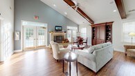 This single-story space is defined by its dramatic vaulted ceilings accented with rich, exposed wood beams, creating an airy and welcoming atmosphere throughout. The main lounge area serves as the heart of the space, centered around a stone fireplace with a decorative mantel and complemented by a wall-mounted television. Comfortable seating, including tufted armchairs and a plush sofa, is arranged to encourage conversation and relaxation, all set atop warm hardwood flooring. Multiple sets of French doors line the walls, flooding the interior with natural light and offering views of the surrounding greenery. A ceiling fan overhead adds to the casual, comfortable ambiance. Adjacent to the lounge, a kitchen area features warm wood cabinetry, a gas cooktop, and a generous counter with an elegantly arched pass-through opening. Bar-height seating at the counter makes it an ideal spot for casual dining or gatherings. A separate room provides a more private setting, furnished with a round dining table and rich wood built-in cabinetry with open shelving, suitable for meetings or office use. Together, these areas create a cohesive and versatile communal environment.