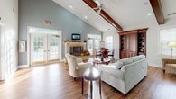 This single-story space is defined by its dramatic vaulted ceilings accented with rich, exposed wood beams, creating an airy and welcoming atmosphere throughout. The main lounge area serves as the heart of the space, centered around a stone fireplace with a decorative mantel and complemented by a wall-mounted television. Comfortable seating, including tufted armchairs and a plush sofa, is arranged to encourage conversation and relaxation, all set atop warm hardwood flooring. Multiple sets of French doors line the walls, flooding the interior with natural light and offering views of the surrounding greenery. A ceiling fan overhead adds to the casual, comfortable ambiance. Adjacent to the lounge, a kitchen area features warm wood cabinetry, a gas cooktop, and a generous counter with an elegantly arched pass-through opening. Bar-height seating at the counter makes it an ideal spot for casual dining or gatherings. A separate room provides a more private setting, furnished with a round dining table and rich wood built-in cabinetry with open shelving, suitable for meetings or office use. Together, these areas create a cohesive and versatile communal environment.