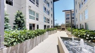 This thoughtfully designed outdoor courtyard offers a refined urban retreat set between two modern multi-story buildings. The space is defined by clean, contemporary architecture featuring smooth concrete finishes, large-format tile paving, and a neutral palette that creates a calm, cohesive aesthetic throughout.

At the heart of the courtyard stands a striking concrete fireplace, flanked by a pair of minimalist wood-and-concrete benches that invite visitors to gather and relax. The fireplace serves as a focal point, anchoring the space with a sense of warmth and community. On either side, raised concrete planters overflow with lush, varied greenery — including ornamental shrubs, tropical foliage, and desert-adapted plants — adding vibrant texture and a connection to nature within the urban setting.

A spacious central walkway runs the length of the courtyard, bordered by the planters on one side and a water feature on the other. The fountain elements introduce a gentle, soothing ambiance to the environment. Overhead, a dramatic architectural bridge connects the two flanking buildings, framing an open view of the bright blue sky and the surrounding cityscape beyond.

The courtyard is bathed in abundant natural light, making it a bright and welcoming open-air space suited for relaxation or casual interaction.