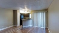 This single-floor space offers a practical and well-appointed open-concept layout that seamlessly connects the living area and kitchen. The living space features light-toned hardwood-style flooring that extends throughout, adding warmth and continuity to the interior. A ceiling fan overhead provides comfort and airflow, while a full-height vertical blind-covered sliding glass door allows natural light to fill the room and provides access to the outdoors.

The kitchen is defined by dark espresso cabinetry that contrasts elegantly against a stone tile backsplash and a granite-style countertop. A raised breakfast bar serves as a natural divider between the kitchen and living area, making it ideal for casual dining or entertaining. Black appliances, including a refrigerator and microwave, complement the kitchen's rich, dark aesthetic. A window above the sink brings additional natural light into the cooking space.

The textured popcorn ceiling is consistent throughout, lending a classic residential character to the unit. A door off the main living area provides access to additional rooms or storage. Overall, the space presents a cohesive design with functional layout choices, durable finishes, and a welcoming atmosphere suited for everyday living.
