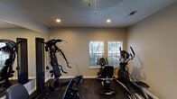Located on W Pleasant Run Rd in Lancaster, Texas, this single-floor fitness space is thoughtfully designed to support a full range of workout routines. The room features dark rubber flooring throughout, providing a durable, slip-resistant surface well-suited for high-intensity training and heavy equipment use.

Natural light flows in generously through multiple windows fitted with horizontal blinds, offering views of the surrounding neighborhood while keeping the space bright and inviting. Recessed ceiling lighting and a ceiling fan supplement the natural light, ensuring consistent illumination throughout the room.

A full wall of floor-to-ceiling mirrors visually expands the space and allows users to monitor their form during workouts. The equipment selection is extensive, including a multi-function cable machine, an elliptical trainer, a stationary recumbent bike, a treadmill, and a dedicated free weights area with a dumbbell rack and medicine ball storage. The neutral, warm-toned walls contribute to a focused yet welcoming atmosphere.

Interior pass-through windows connect the fitness room to an adjacent area, adding a sense of openness. The space is well-ventilated with visible HVAC vents, making it functional and comfortable for sustained exercise sessions.