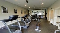 This fitness center is thoughtfully designed to support a full range of workout routines in a comfortable, single-floor setting. The space features a dedicated cardio zone with treadmills and an elliptical machine, providing ample equipment for aerobic exercise. A multi-station cable and weight machine anchors the strength training area, complemented by weight benches positioned for functional use throughout the room. The flooring consists of durable rubber gym matting, providing safety and stability during workouts. Recessed lighting is evenly distributed across the ceiling, ensuring the space is well-lit throughout. Ceiling fans contribute to air circulation, enhancing comfort during intense training sessions. A large wall-mounted mirror spans a significant portion of one wall, visually expanding the space and allowing users to monitor their form. Motivational framed posters add a purposeful aesthetic to the walls, reinforcing the fitness-focused atmosphere. The overall ambiance is clean, functional, and professional, making this fitness center a practical and inviting space for a broad range of exercise needs.