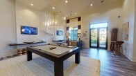This well-appointed community clubroom is designed for both leisure and socializing, showcasing a thoughtfully curated interior with soaring ceilings that lend an open, airy atmosphere throughout. The main area is anchored by a full-size billiards table beneath a striking geometric gold chandelier, complemented by recessed lighting that illuminates the warm, neutral-toned hardwood-style flooring. Shiplap-accented walls add a modern-coastal character, while a wall-mounted flat-screen TV and a built-in media console provide entertainment options for guests. A dedicated lounge nook features plush seating, a coffee table, and floor-to-ceiling windows dressed in bold brown-and-white striped curtains, flooding the space with natural light and offering views of the lush outdoor surroundings. French doors lead directly to an exterior courtyard, seamlessly blending indoor and outdoor living. A casual dining area with a round wood table and upholstered chairs is accented by a wall-mounted cue rack and an eye-catching collection of organic driftwood art panels. A high-top bar table near the entry provides an additional social spot, while a media and game storage unit rounds out the amenities, making this clubroom a versatile and inviting gathering space.