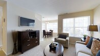 The Mill Ridge Model is a thoughtfully designed single-story residence that showcases a seamless, open-concept layout connecting the living and dining areas. The living space is carpeted and centered around a wall-mounted flat-screen television positioned above a dark-stained media console with ample storage drawers, adding a rich contrast to the neutral, light-toned walls. A round coffee table anchors the seating arrangement, complemented by a plush armchair with accent pillow, creating a comfortable and functional gathering area.

The transition into the dining area is natural and uninterrupted, where a round dining table surrounded by upholstered chairs sits beneath an elegant chandelier. This area benefits from an abundance of natural light, courtesy of large windows fitted with horizontal blinds that line multiple walls, offering bright, airy views of the outdoors. The consistent use of light-toned walls and neutral flooring throughout reinforces a cohesive, welcoming aesthetic. Overall, the Mill Ridge Model presents a well-balanced combination of functional design and inviting ambiance, ideal for comfortable everyday living.