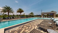 This outdoor amenity area centers around a generously sized swimming pool with clear turquoise water, designed in a classic rectangular layout with a gradual depth progression clearly marked along the pool's edge. The pool deck is finished in a distinctive multi-toned interlocking paver pattern, combining warm beige, tan, and deep brown tones to create a visually striking surface that extends broadly around the pool on all sides.

The surrounding deck is well-furnished with numerous slatted lounge chairs paired with light blue cushions, arranged in organized rows along the poolside. Coordinating navy blue market umbrellas are positioned throughout the space, providing ample shade for guests. Additional seating areas are distributed across the deck, including clustered conversational groupings with cushioned chairs and low tables, ideal for relaxed outdoor gatherings.

Tall, mature palm trees are planted throughout the area, adding a tropical character and natural shade. A covered pavilion structure with a pitched roof anchors one end of the deck, offering a shaded retreat. Wrought iron fencing encloses the perimeter, beyond which open green landscape and distant treelines are visible, enhancing the sense of openness. The overall ambiance is bright, airy, and resort-inspired.