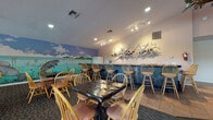 Carlton Arms in Ocala, Florida offers a richly decorated single-floor community clubhouse designed to inspire and entertain. The main gathering area features soaring vaulted ceilings with recessed lighting and large windows that frame outdoor views, flooding the space with natural light. Seating is plentiful, with a mix of upholstered lounge chairs, Windsor-style dining chairs, and small tables arranged across patterned carpet and tile flooring.

The space is defined by its striking hand-painted murals. A sweeping equestrian scene stretches across one wall, depicting horses grazing along a fenced Florida pasture — a nod to Ocala's renowned horse country heritage. A second, equally captivating mural depicts Florida's native manatees in a vibrant underwater and coastal scene, complete with clouds and sea grass.

Suspended decorative fish sculptures add a whimsical, nature-inspired touch overhead, while an intricate driftwood wall installation anchors the bar area with artistic flair. The rustic bar features a dark wood counter with bar stool seating and is equipped with a full-size refrigerator and basic kitchen amenities — ideal for community gatherings.

A cozy lounge area with a large flat-screen TV and a distinctive ship's wheel glass-top coffee table provides a comfortable retreat. Throughout, the space blends Florida's natural and equestrian themes into a warm, welcoming community environment.