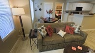 This single-floor residence offers an open-concept layout that seamlessly connects the living, dining, and kitchen areas, creating a cohesive and functional living environment. The living room serves as the heart of the space, anchored by a plush olive-green sofa adorned with a mix of patterned and textured throw pillows in rich earth tones of red, brown, and cream. Light hardwood-style flooring runs throughout, adding warmth and continuity across the open floor plan. A compact side table and floor lamp provide practical accents to the seating area. Adjacent to the living room, a dark-topped dining table with modern white chairs offers an intimate setting for meals, complemented by a decorative floral centerpiece that adds a pop of color. The wall décor, featuring an arrangement of round mirrors and plates, adds artistic character to the space. Beyond the dining area, a glimpse into the bedroom reveals a neatly appointed sleeping space with natural light filtering through window blinds. The kitchen, visible from the main living area, features white cabinetry and offers convenient accessibility. Neutral, warm-toned walls tie the entire space together, creating a comfortable and welcoming ambiance throughout.