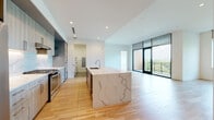 This single-floor residence showcases a contemporary open-concept design with clean lines, recessed lighting, and light-toned hardwood flooring that flows throughout the main living areas. The kitchen serves as a central focal point, anchored by a substantial waterfall-edge island finished in dramatic veined stone. Cabinetry features a refined wood-grain finish, complemented by a matching stone backsplash, stainless steel appliances including a gas range, built-in ovens, and a side-by-side refrigerator with water dispenser.

The expansive living and dining areas benefit from abundant natural light through floor-to-ceiling glass doors that frame treetop views, creating a seamless indoor-outdoor connection. A built-in shelving nook adds functional detail to the open layout.

Bedrooms are carpeted for comfort and feature large windows with exterior views and ceiling fans. A generously sized walk-in closet with built-in shelving and hanging rods provides ample storage.

Bathrooms include a full bath with tub and wood-tone cabinetry, and a stylish half bath with a statement geometric-patterned accent wall and a floating walnut vanity. A dedicated laundry room houses a full-size washer and dryer with overhead shelving.