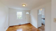This two-floor home presents a clean, straightforward layout with consistent design elements carried throughout both levels. Warm-toned hardwood flooring runs through the main living areas and bedrooms, lending a cohesive and inviting character to the interior. Neutral white walls keep the spaces feeling bright and adaptable.

The main level features a welcoming entry with a paneled front door topped by a arched transom window, allowing natural light to filter into the foyer. Adjacent to the entry, the living area is open and well-lit, with a large window offering views of the surrounding neighborhood. The kitchen is tucked to the side and includes white cabinetry, dark laminate countertops, a stainless steel sink, and standard appliances including a refrigerator and dishwasher. A window above the sink provides natural light to the workspace. Tile flooring grounds the kitchen area.

A staircase with a wooden handrail connects to the upper level, where multiple bedrooms are found. Each bedroom features the same warm hardwood flooring and windows that bring in ample natural light with views of greenery outside. The bathroom on the upper level showcases retro-style yellow square tiles with black accent trim, a full soaking tub, and a window for ventilation.