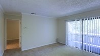 This single-floor residence offers a clean and functional layout with a calm, neutral aesthetic throughout. The main living area is finished with wall-to-wall carpeting in a warm beige tone, providing a comfortable and inviting atmosphere underfoot. Smooth crown molding frames the junction between the walls and the textured popcorn ceiling, adding a subtle architectural detail to the space. The room is bathed in natural light, thanks to a large sliding glass door fitted with vertical blinds that can be adjusted to control light and privacy. The door opens to a verdant outdoor setting, with visible foliage creating a pleasant connection to the outside. A wall-mounted thermostat and a floor-level air vent indicate a central HVAC system for year-round climate control. The walls are painted in a soft, neutral tone that complements the light carpeting and contributes to the bright, airy feel of the interior. Overall, the space presents a straightforward and well-maintained single-level layout suitable for comfortable everyday living.